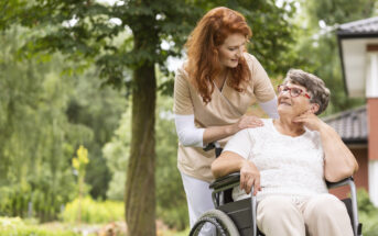 A caregiver with red hair smiles and leans over a seated elderly woman in a wheelchair. The elderly woman, wearing glasses and a white shirt, looks up at the caregiver. They are outside in a garden setting with trees and a building in the background.