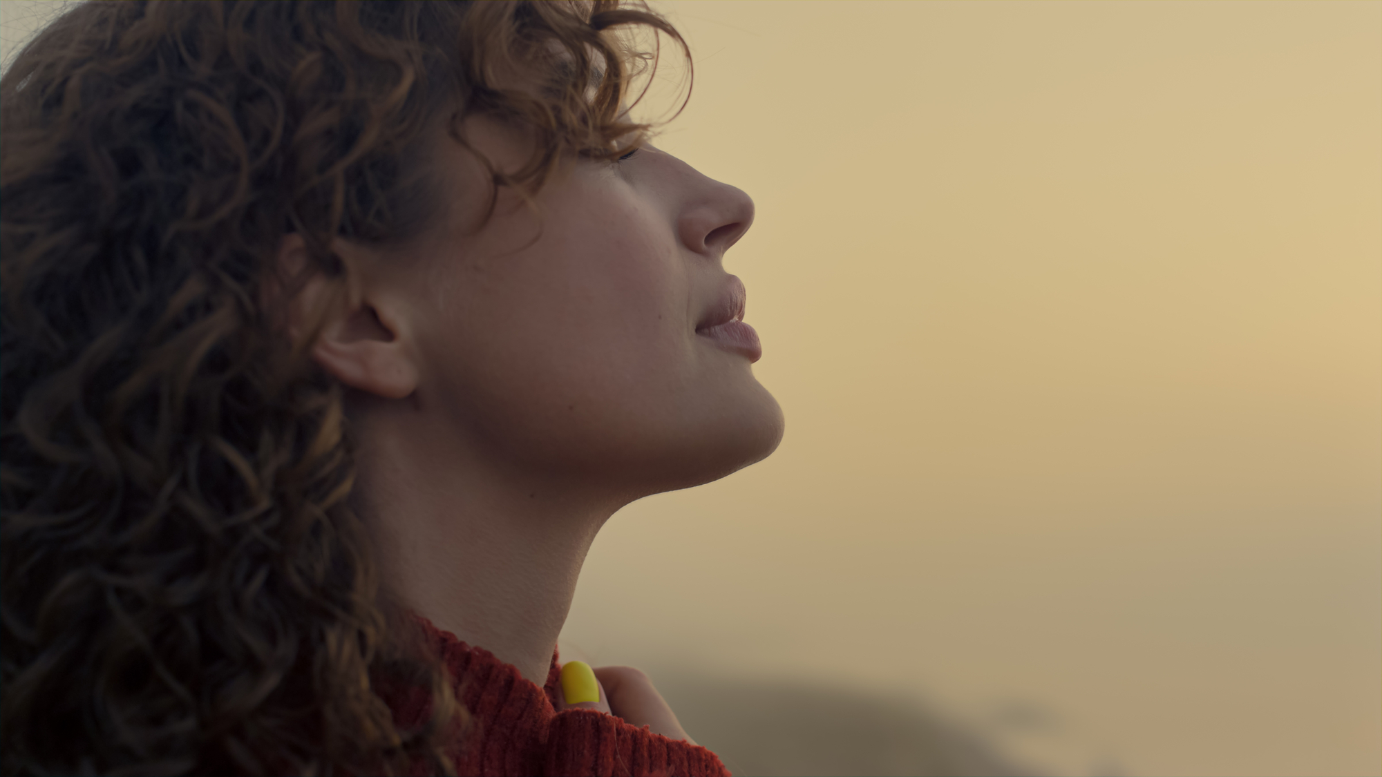 A person with curly hair is shown in a close-up side profile against a hazy, light background. They are wearing a red sweater and looking upward with a serene expression. The soft light creates a calm and contemplative atmosphere.