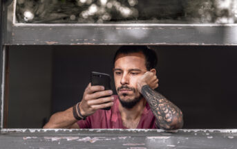 A man with a beard and a sleeve tattoo leans on a windowsill, looking intently at his smartphone. He is wearing a maroon T-shirt and has his hair tied back. The window frame is old and worn, and there are blurred white lights in the background.