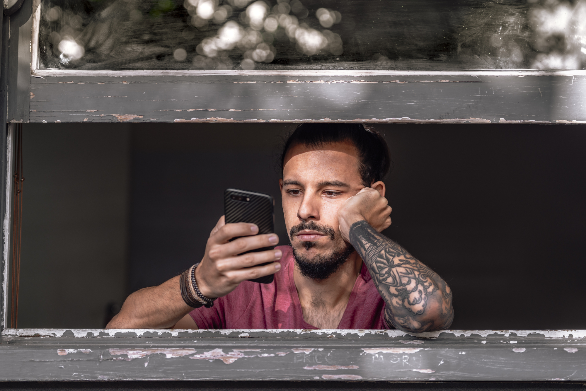 A man with a beard and a sleeve tattoo leans on a windowsill, looking intently at his smartphone. He is wearing a maroon T-shirt and has his hair tied back. The window frame is old and worn, and there are blurred white lights in the background.