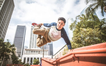 A man dressed in a blue and white long-sleeve shirt and beige pants performs a parkour move by jumping over a red wall in an urban environment. Tall buildings and trees can be seen in the background, suggesting a city setting.