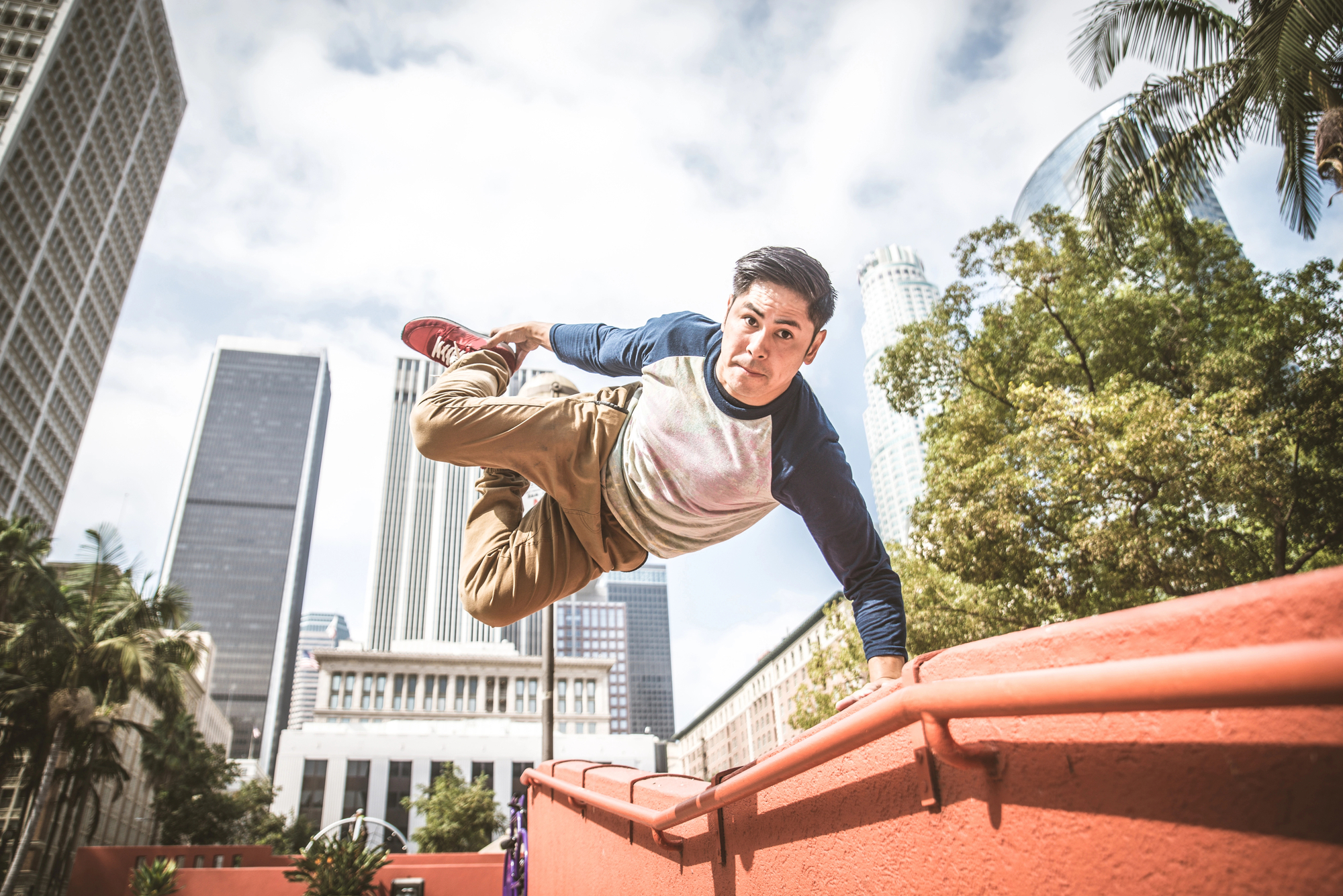 A man dressed in a blue and white long-sleeve shirt and beige pants performs a parkour move by jumping over a red wall in an urban environment. Tall buildings and trees can be seen in the background, suggesting a city setting.