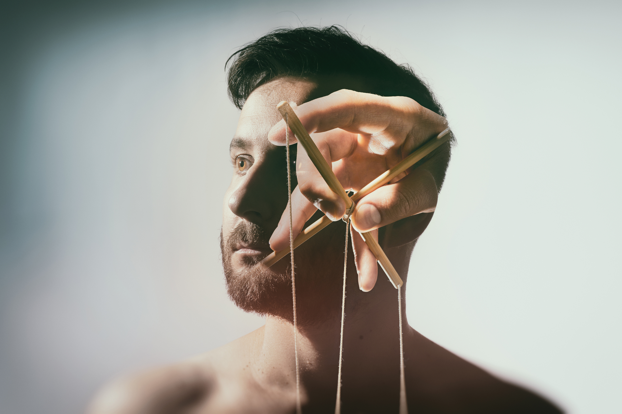 A man with a serious expression is depicted with a large hand holding marionette strings in front of his face, making him appear controlled like a puppet. The image combines elements of portraiture and surrealism, suggesting themes of manipulation and control.