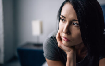 A woman with short dark hair is resting her face on her hand, looking thoughtfully into the distance. She appears pensive and is sitting indoors with a blurred background of furniture. She is wearing a gray top.