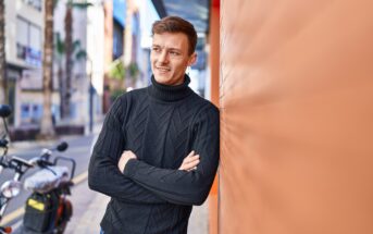 A man wearing a black turtleneck sweater stands with his arms crossed and leans against an orange wall. He is smiling and looking off into the distance. A motorbike and palm trees line the street in the background. The scene is sunny and urban.