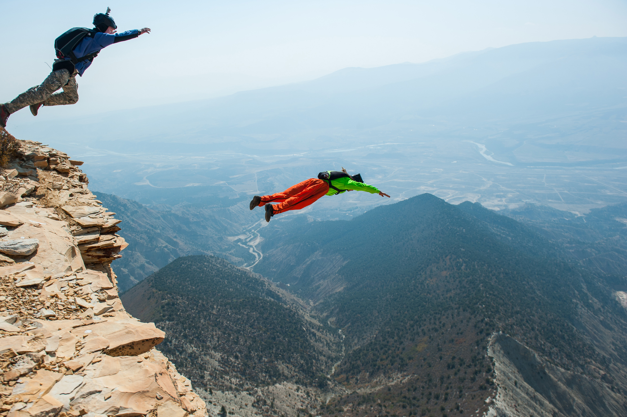 Two people wearing wingsuits leap off a rocky cliff into a vast mountainous landscape. The sky is hazy, and the valley below is a mix of greenery and barren land. The image captures the thrill and adventure of BASE jumping in nature.