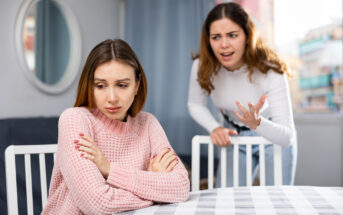 Two women are in a tense situation at a table. The woman on the left, wearing a pink sweater, sits with arms crossed, looking upset. The woman on the right, in a white top, stands behind her with an expressive, frustrated gesture, appearing to speak intensely.