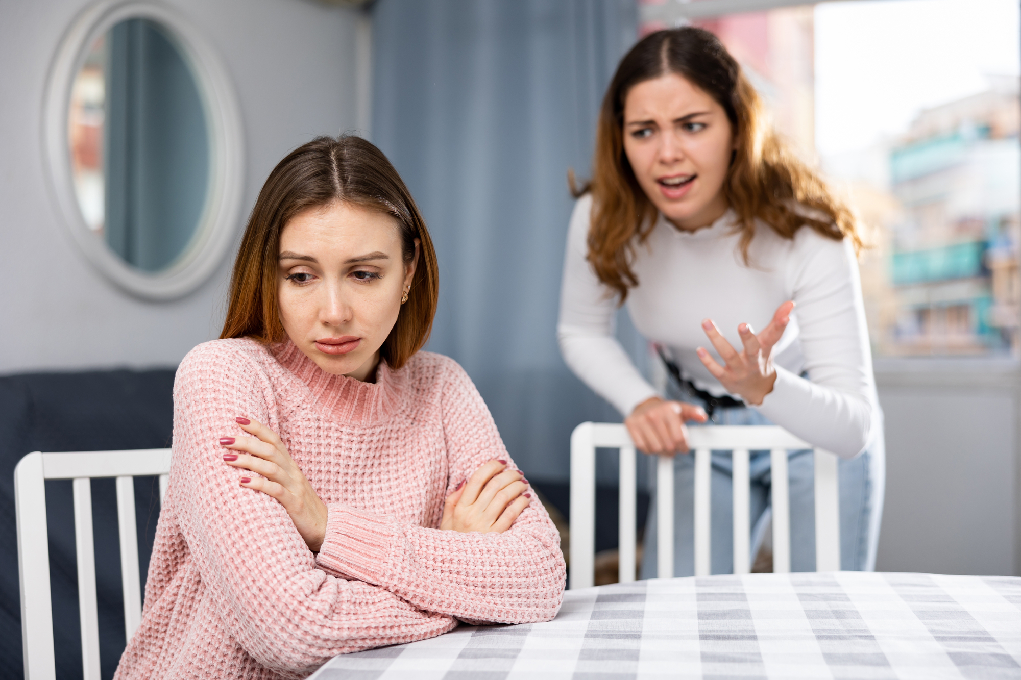 Two women are in a tense situation at a table. The woman on the left, wearing a pink sweater, sits with arms crossed, looking upset. The woman on the right, in a white top, stands behind her with an expressive, frustrated gesture, appearing to speak intensely.