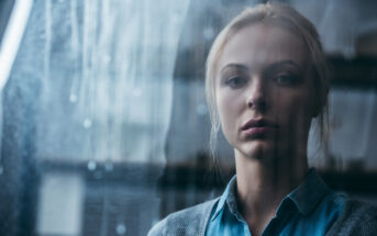 A woman with blonde hair is standing behind a rain-streaked window. She is wearing a blue shirt and a gray cardigan, looking pensively into the distance. The background is blurred and out of focus.