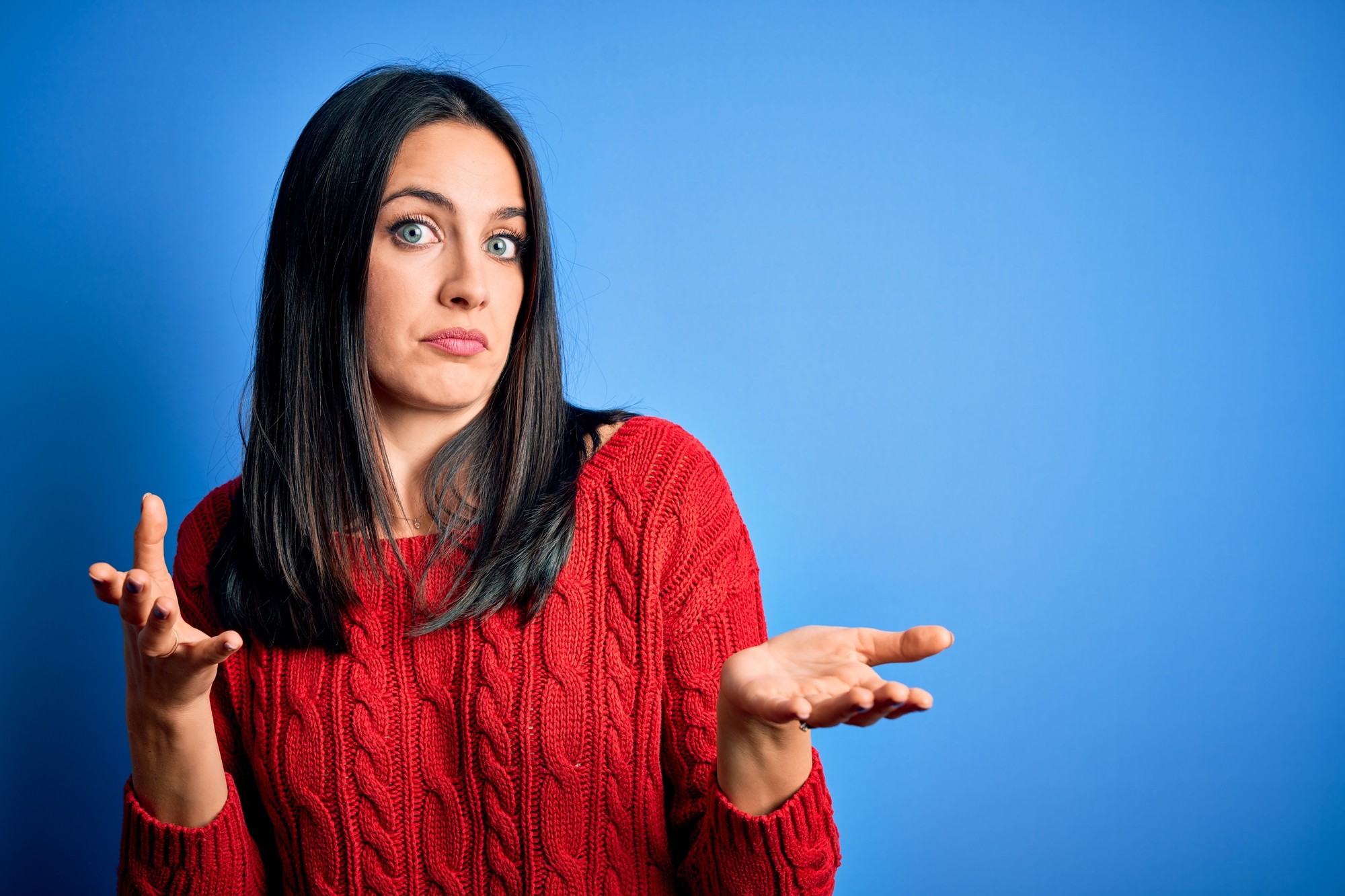 A person with long dark hair wearing a red knit sweater stands against a blue background. They are looking forward with their eyebrows slightly raised and eyes wide open, and they have their arms out to the sides with their palms up in a questioning or confused gesture.