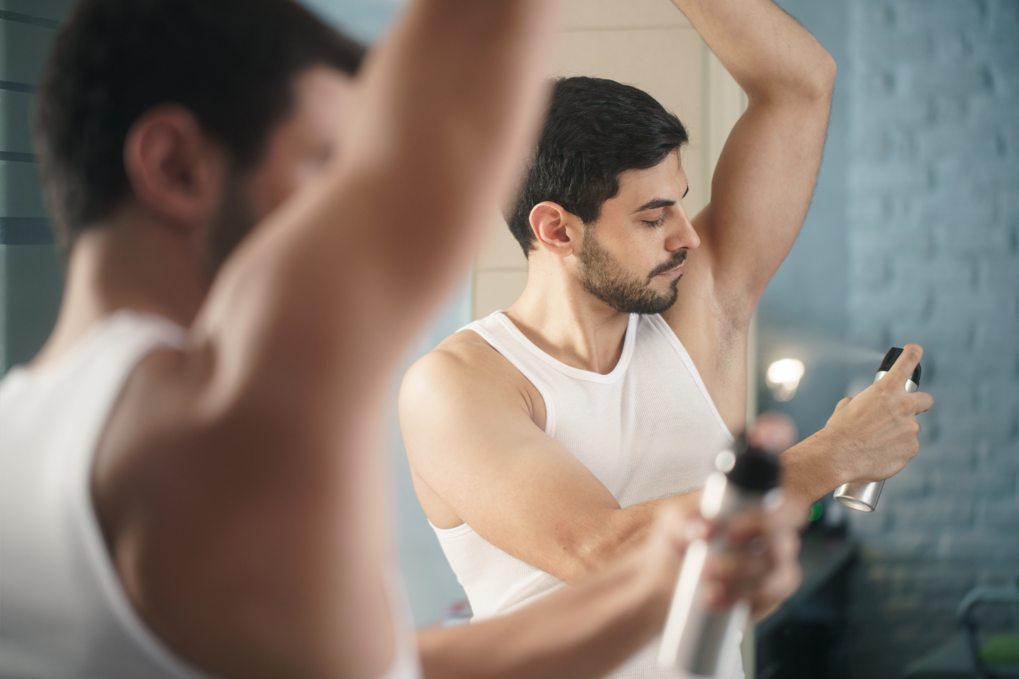 A man is standing in front of a mirror, wearing a white tank top, and applying deodorant to his underarm with his right arm raised. His reflection is visible in the mirror, showing his focused expression. The background includes a blurred bathroom setting.