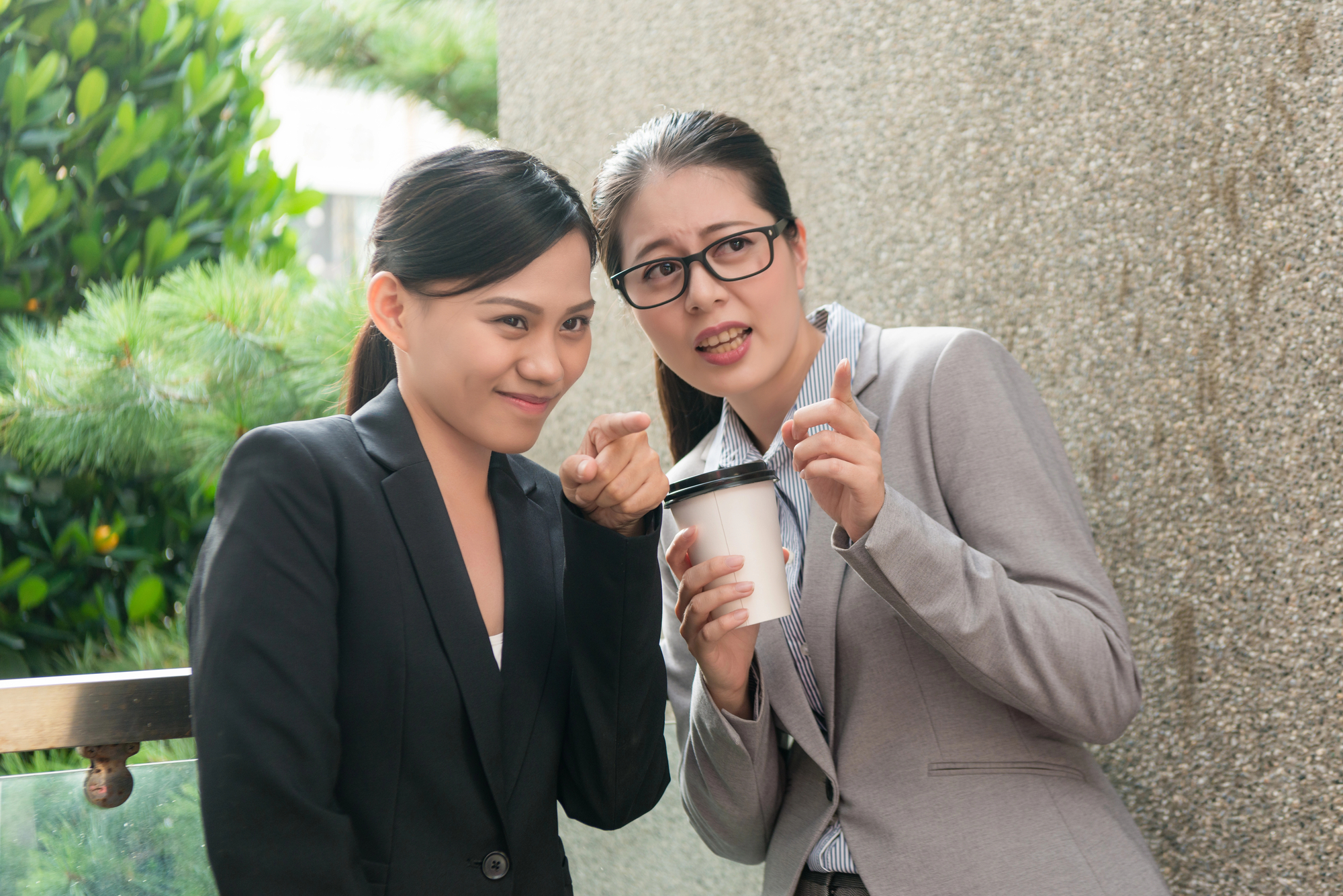 Two women dressed in business attire stand close together. One woman points at the camera and smiles, while the other, who wears glasses, holds a disposable coffee cup and points as well. They appear to be outside against a wall with greenery in the background.