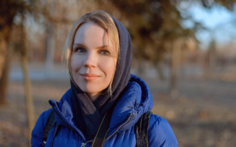 A woman wearing a blue jacket and a dark hood stands outdoors. She has a slight smile and blonde hair peeking out from her hood. The background is blurred, featuring trees and a soft light creating a peaceful atmosphere.