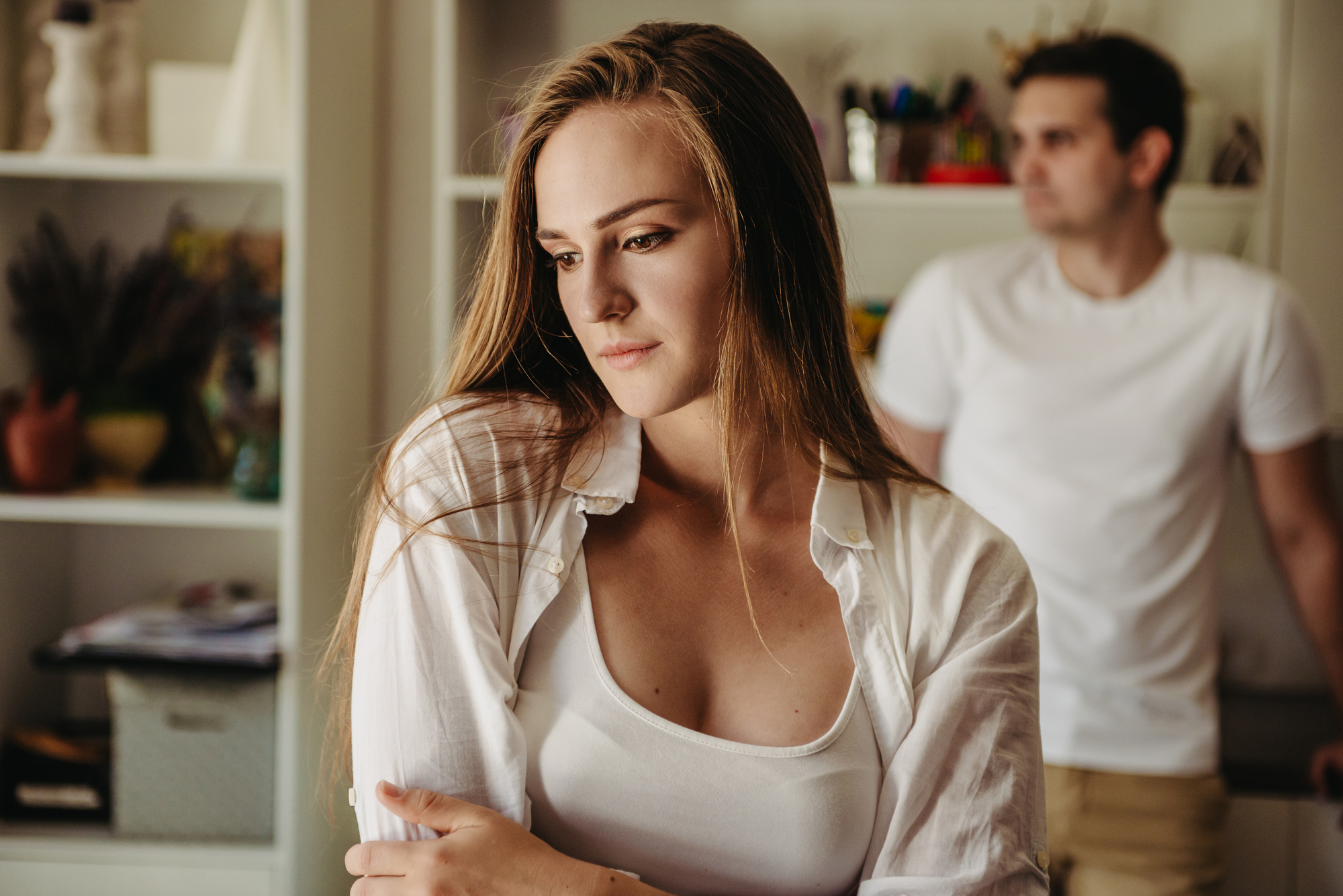 A woman with long hair and a white shirt stands in the foreground, looking contemplative, while a man in a white shirt stands in the blurred background. They are inside a room with shelves filled with various items.