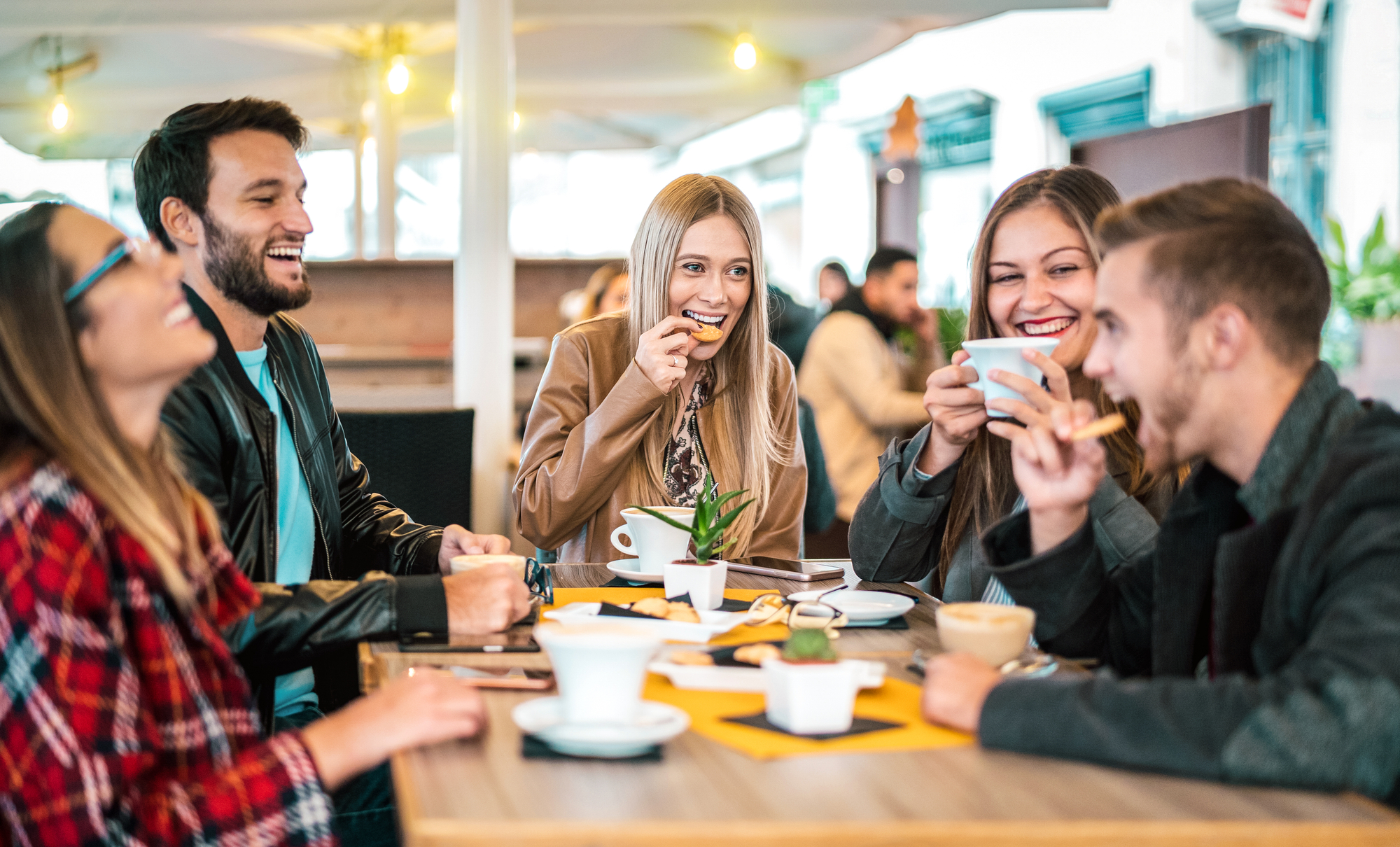 A group of five friends is sitting around a table at an outdoor cafe. They are smiling and laughing while enjoying coffee and snacks. The atmosphere appears warm and convivial, with people interacting cheerfully.