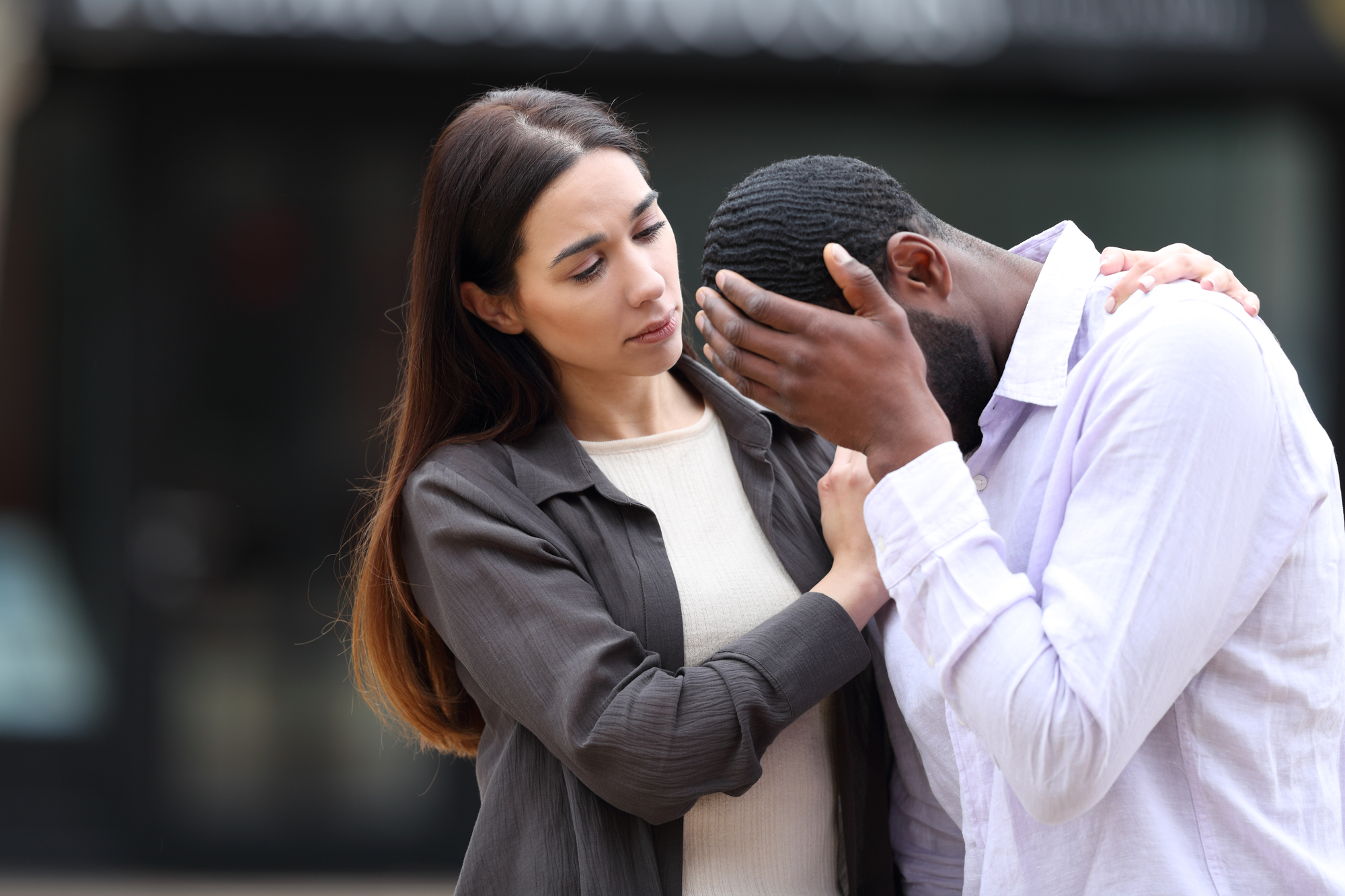 A woman with long hair comforts a man who is visibly upset. She has one arm around his shoulder and her other hand on his arm. He is holding his head in his hands. Both are outdoors, and the background is blurred, focusing the attention on the pair.