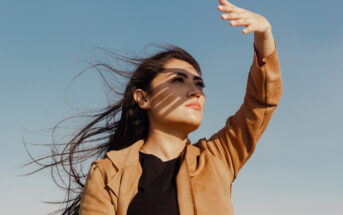 A woman with long hair, wearing a brown jacket and black shirt, raises her hand to shield her eyes from the sun. Shadow patterns cross her face as she looks upwards with a serene expression. The blue sky in the background emphasizes the bright, sunny day.