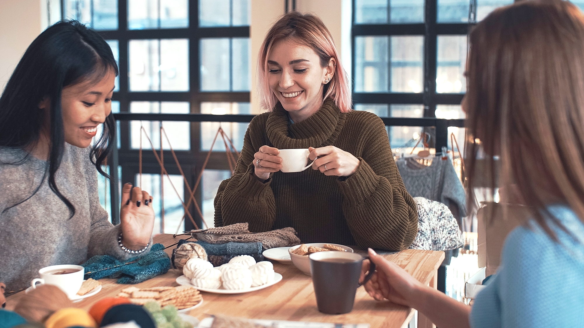 Three women sit around a wooden table enjoying hot beverages and snacks. One woman, with pink hair, holds a cup and smiles. Knit items and yarn are scattered on the table. Large windows in the background let in natural light, creating a cozy atmosphere.