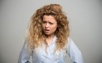 A woman with long, curly hair and a light blue shirt stands against a light gray background. She has a slightly furrowed brow and a puzzled or questioning expression.