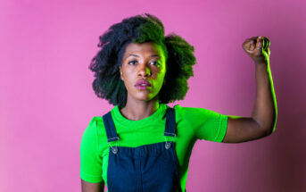 A person with curly hair, wearing a bright green shirt and denim overalls, stands against a pink background. They raise their right arm with a clenched fist in a gesture of strength and determination.