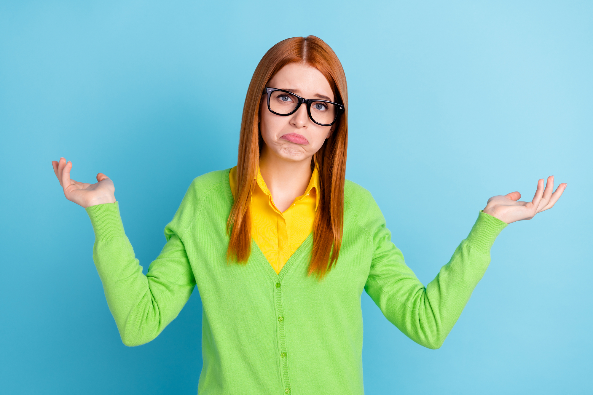 A person with long red hair, wearing black glasses, a bright green cardigan, and a yellow shirt stands against a blue background, shrugging with a puzzled expression on their face and raising both hands with palms upwards.