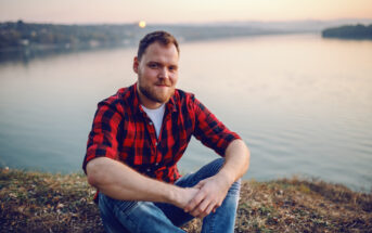 A man with a beard, wearing a red and black plaid shirt and blue jeans, sits on the grass by a calm lake during sunset. The background features a serene body of water, distant hills, and a soft, pastel sky. He is smiling gently at the camera.