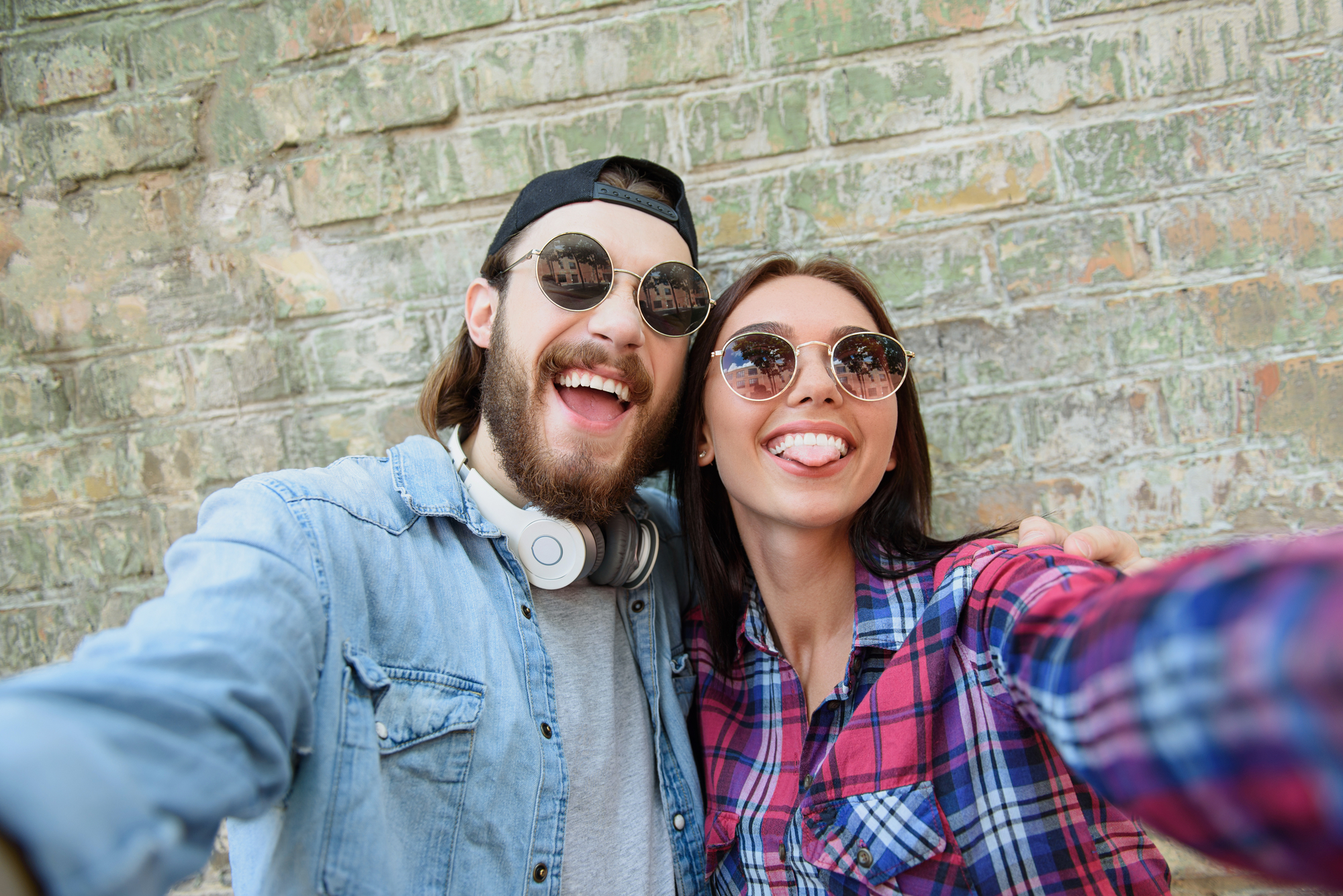 A bearded man and a woman, both wearing sunglasses, smile and stick out their tongues while taking a selfie. The man wears a black cap, headphones, and a denim shirt. The woman is in a plaid shirt. They are standing in front of a weathered brick wall.