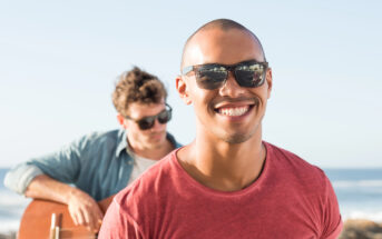 Two men enjoying a sunny day by the beach. The man in the foreground is smiling and wearing sunglasses and a red shirt. The man in the background, also wearing sunglasses, is playing a guitar. The ocean and clear sky are visible.