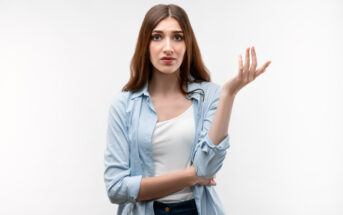 A young woman with long brown hair wearing a light blue button-up shirt over a white top is standing against a plain white background. She looks puzzled or confused, with one arm crossed and the other hand raised with her palm up.