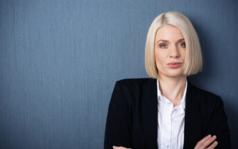 A woman with straight blonde hair cut into a bob stands against a textured blue wall. She is wearing a white collared shirt and a black blazer, with her arms crossed, gazing directly at the camera with a neutral expression.