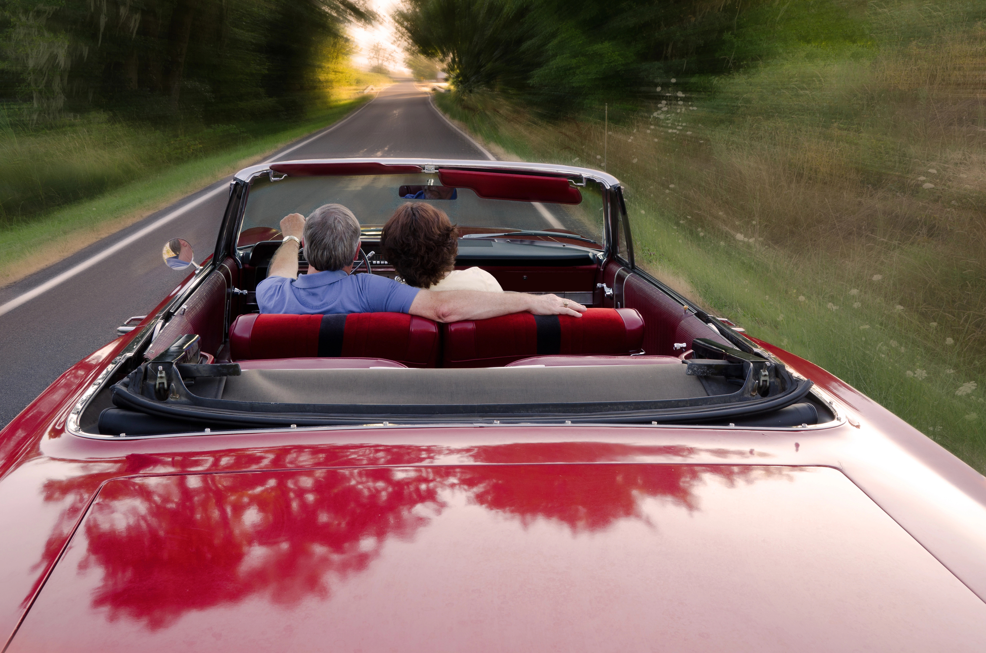 A couple with gray hair enjoys a drive in a red convertible along a tree-lined road. The man drives while the woman leans against him, holding his arm. The car's top is down, capturing a scenic and serene journey.