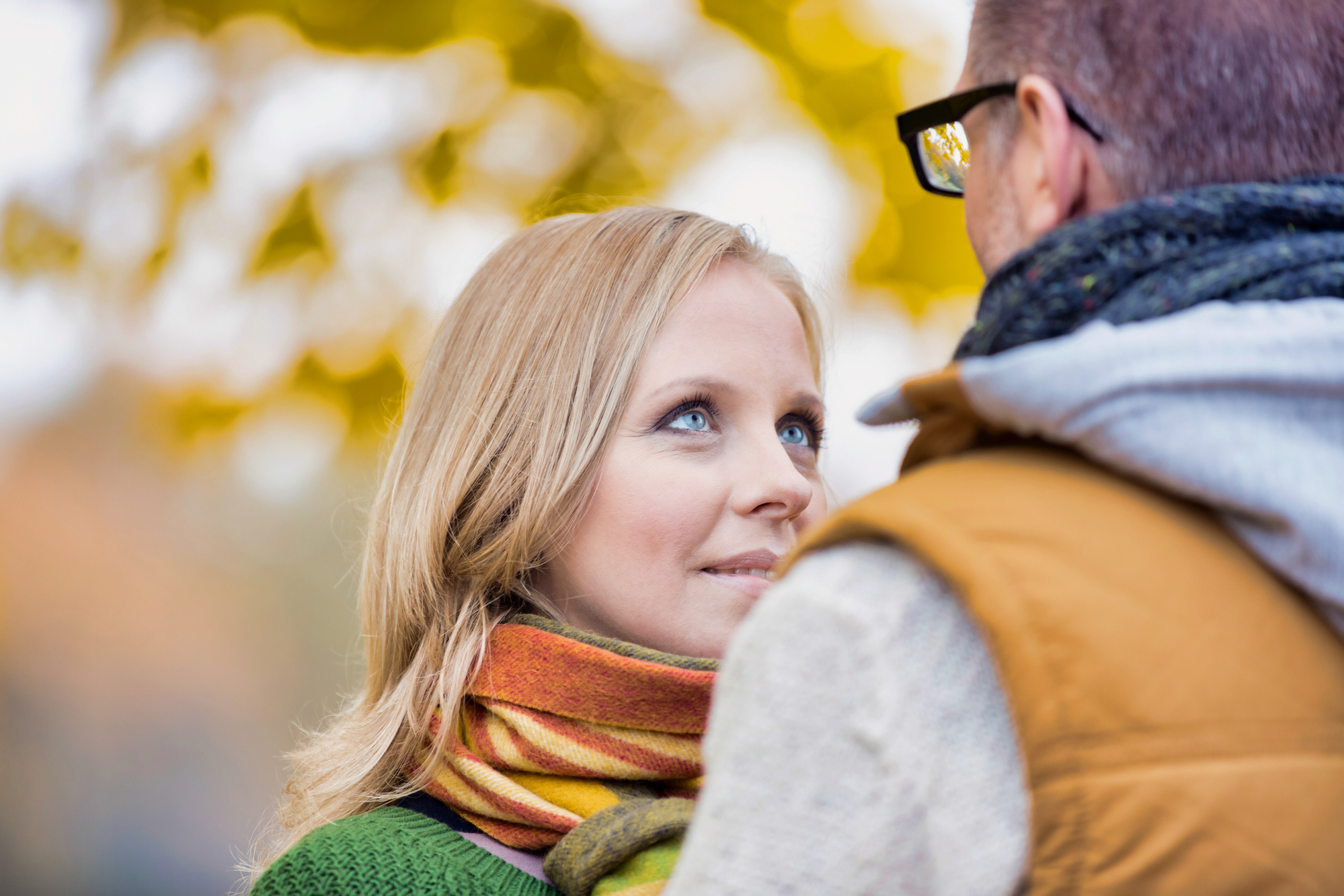 A woman with blonde hair and a colorful scarf looks lovingly into the eyes of a man wearing glasses, a gray hoodie, and a brown vest. They are outdoors, with autumn leaves blurred in the background, creating a warm, romantic setting.