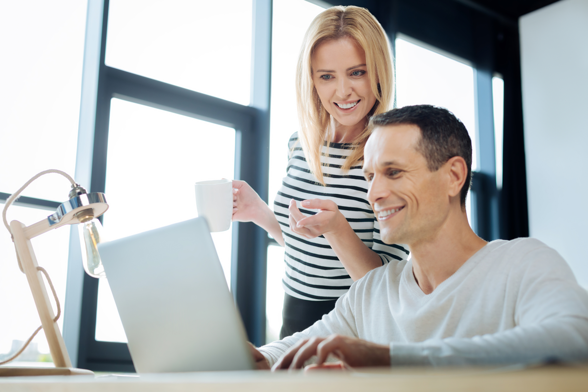 A woman holding a coffee mug stands beside a man who is seated and working on a laptop. They are both smiling and appear to be in a modern, bright office with large windows in the background. The atmosphere is collaborative and friendly.