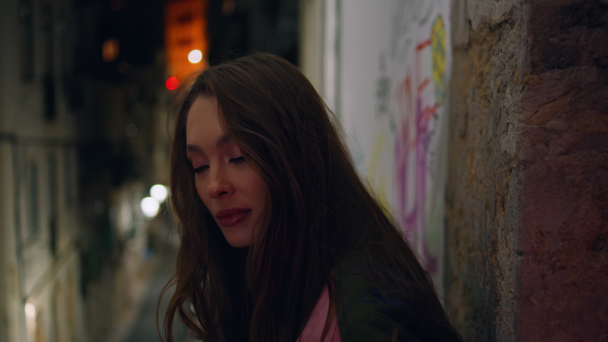 A woman with long hair leans against a graffiti-covered wall in a dimly lit street at night. Her eyes are closed, and her expression is serene. Blurred city lights and another person are visible in the background.