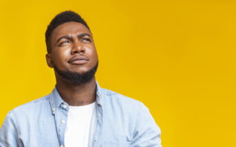 A man with a thoughtful expression looks upward while standing against a bright yellow background. He is wearing a light blue denim shirt over a white t-shirt.