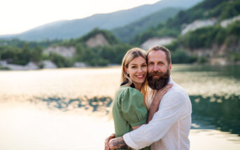 A couple stands by a serene lake with mountains in the background. The woman, wearing a green dress, smiles and rests her hand on the chest of the man with a beard and white shirt. Both look at the camera, enjoying the peaceful, sunset-lit scene.