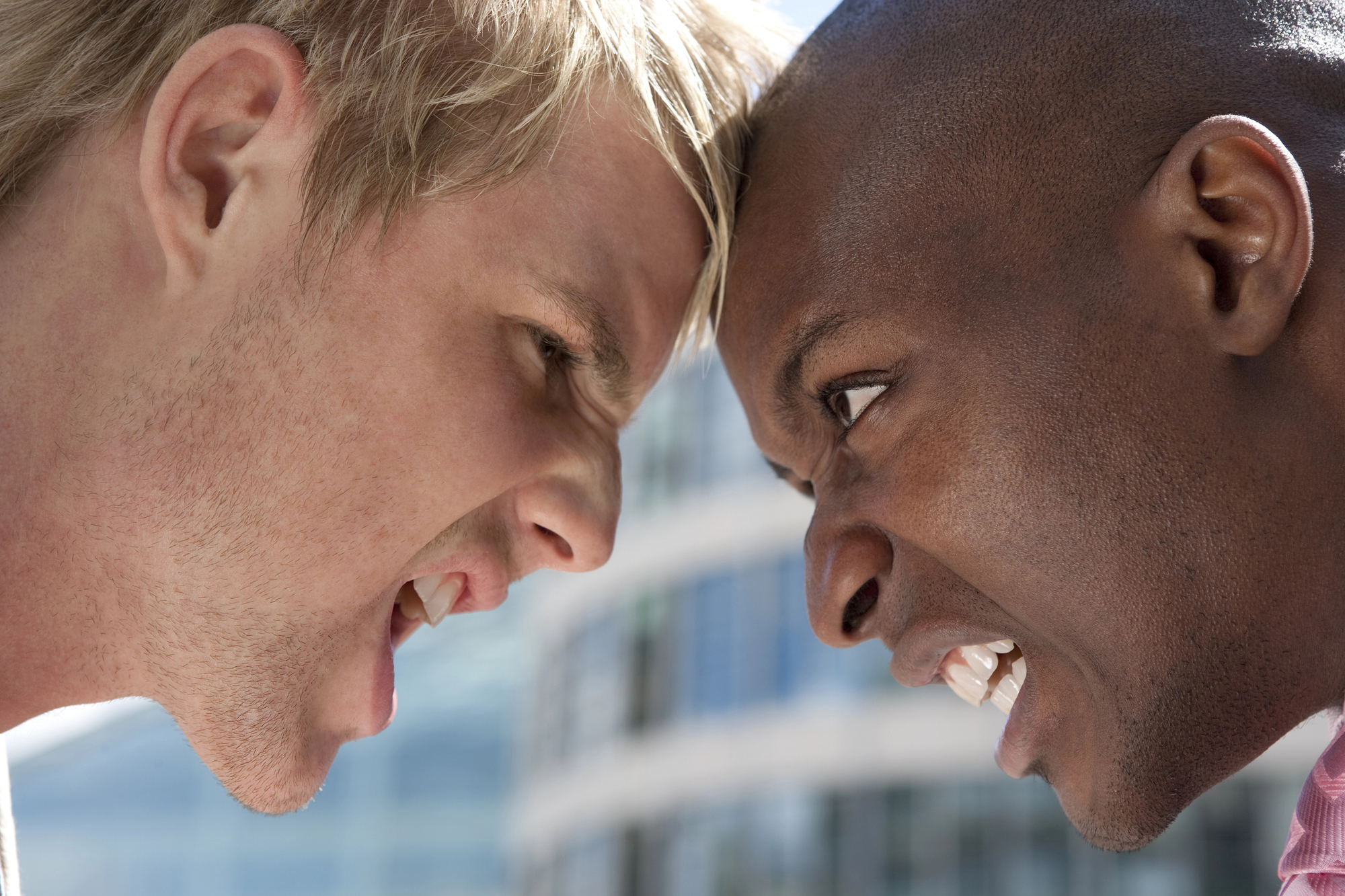 Two men are facing each other with their foreheads touching and angry expressions on their faces. The man on the left has blonde hair and fair skin, while the man on the right has dark skin and a shaved head. They appear to be in a heated confrontation outdoors.