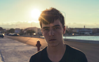 A young person with short, dark hair is standing on an outdoor street at sunset. The sun is low in the sky, casting a warm glow and lens flare. Buildings and a body of water are visible in the background. The person is wearing a dark shirt and has a serious expression.