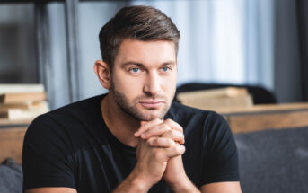 A man with short brown hair and a beard sits with his hands clasped in front of his face. He is wearing a black t-shirt and looking slightly to the left. Books and shelves are visible in the blurred background.