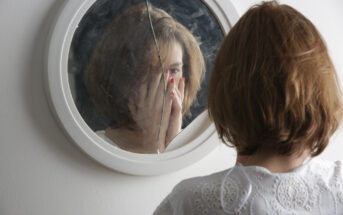 A person with short, brown hair is standing in front of a cracked round mirror, looking at their reflection. They are wearing a white shirt with intricate patterns, and their hand is partially covering their face. The background is a plain white wall.