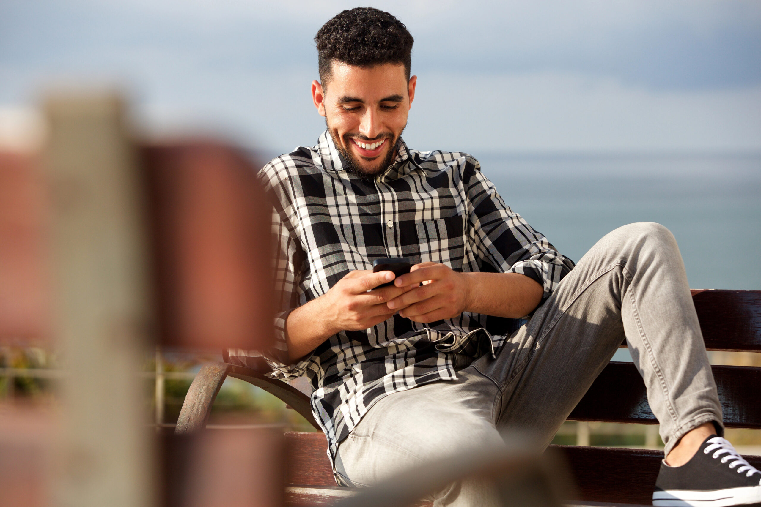 A young man with a beard, wearing a black and white plaid shirt and gray jeans, is sitting on a bench and smiling while looking at his phone. The background includes a blurred view of the ocean and the sky.