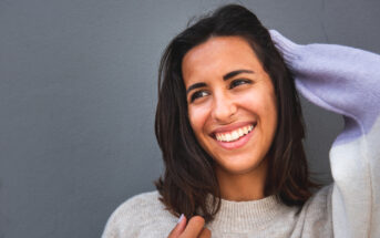 A young woman with shoulder-length dark hair smiles warmly while holding one hand up to her head. She is wearing a light-colored sweater and stands against a plain gray background.