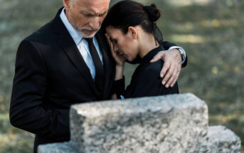 A man and woman, both dressed in black, stand close together at a cemetery. The man, with white hair, embraces the woman, providing comfort. The woman rests her head on the man's chest, appearing distraught. In the foreground, a blurred grave marker is visible.