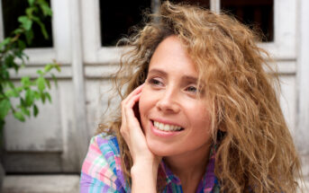 A woman with curly hair smiles while resting her head on her hand. She is wearing a plaid shirt. In the background, there are white doors and some greenery.