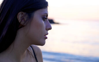 A profile view of a woman with long dark hair looking contemplatively towards the sea at sunset. The calm ocean and gentle colors of the sunset create a serene and introspective atmosphere.