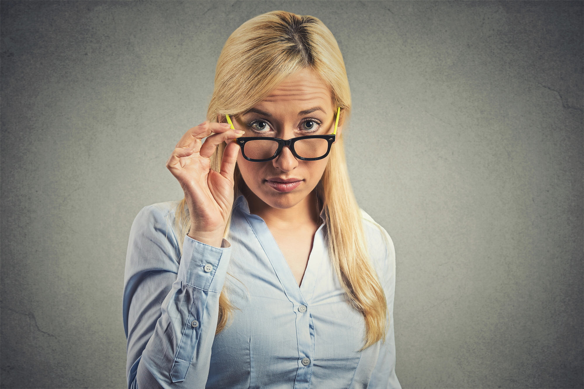 A blonde woman with long hair is standing against a grey background, wearing a light blue button-up shirt. She is looking directly at the camera while adjusting her black-rimmed glasses with one hand, giving an inquisitive expression.