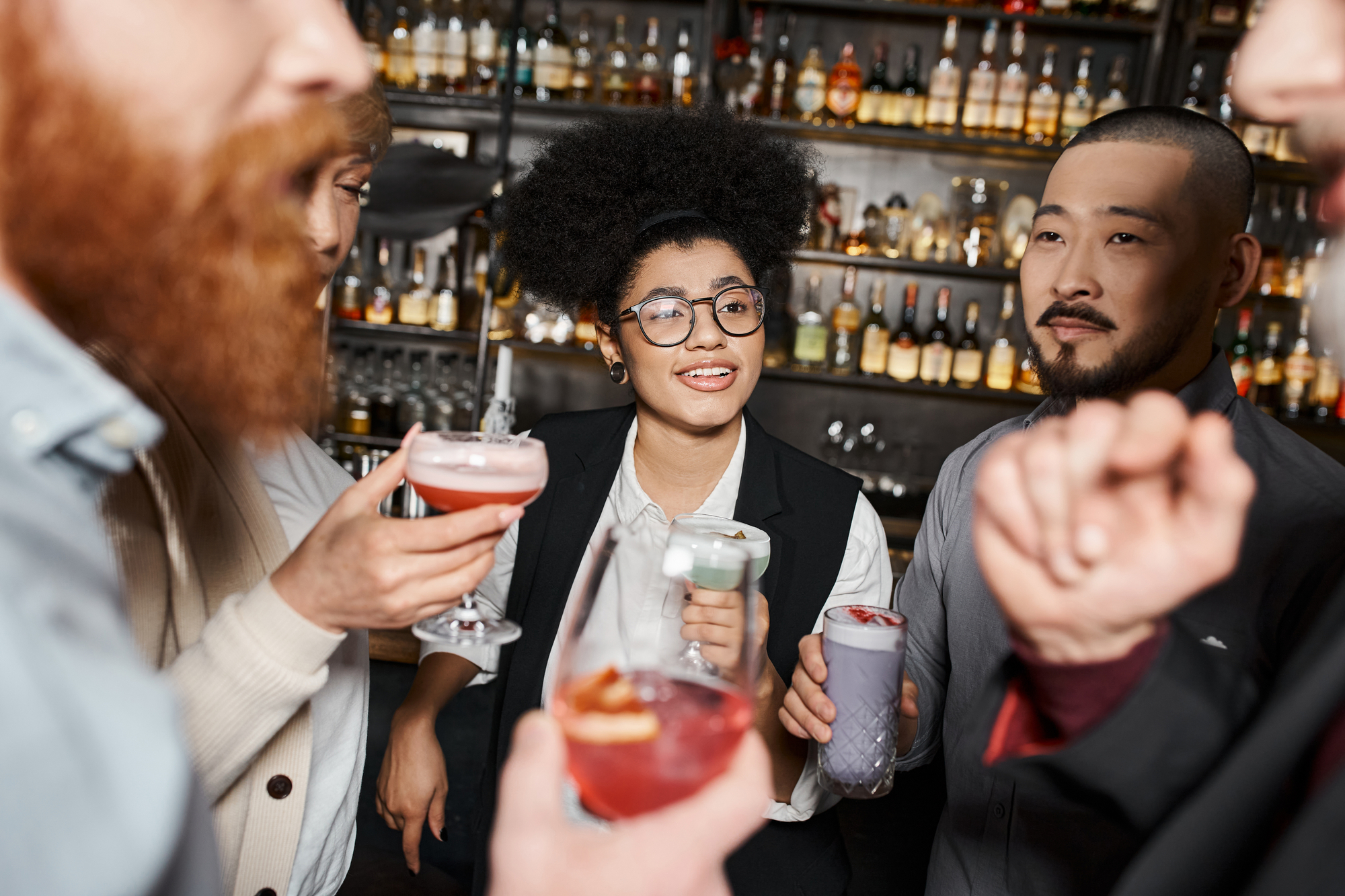 A group of people socializing at a bar. They are holding various cocktails and drinks. The background is filled with shelves stocked with liquor bottles, contributing to a lively atmosphere. Everyone appears to be engaged in conversation and enjoying themselves.