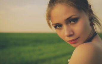 A young woman with light brown hair pulled back gazes softly towards the camera. She is outdoors with a blurred background of a green field and a soft, cloudy sky. She wears subtle makeup and earrings, with one shoulder exposed in the warm light.