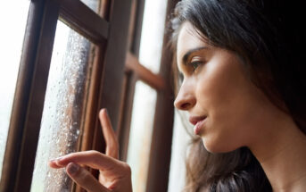 A woman with long dark hair gazes out a window, gently touching the pane with one finger. Raindrops are visible on the glass, and the lighting suggests a natural, soft, and contemplative atmosphere.