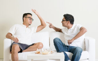 Two men sitting on a white couch engaging in a lively conversation. Both men wear glasses and casual white shirts; one is wearing shorts and the other jeans. The man on the left has his arm raised, while the man on the right points animatedly. A table with snacks sits in front of them.